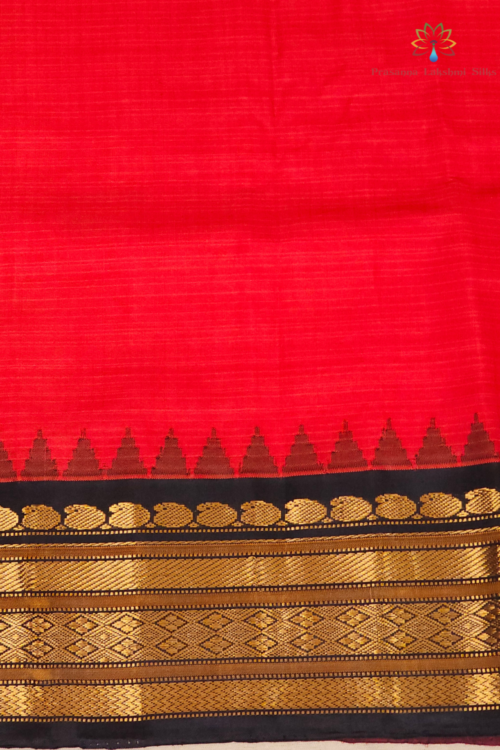 A close-up of a red fabric, likely a saree, featuring a black border with metallic gold zari woven patterns and a row of small gold triangles.